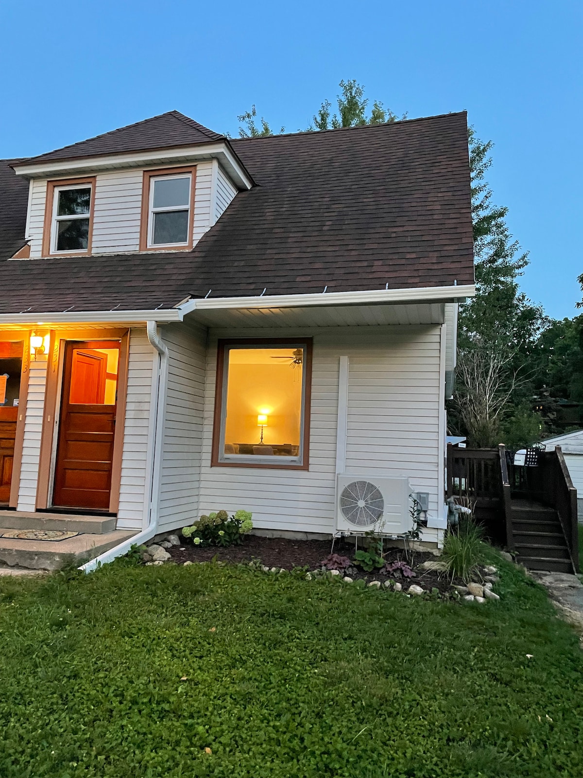 The exterior of the home features a well-maintained façade with light-colored siding and a steeply pitched roof. A warm glow emanates from a window, providing an inviting atmosphere. Lush greenery and decorative stones surround the base of the house, adding natural elements to the setting.