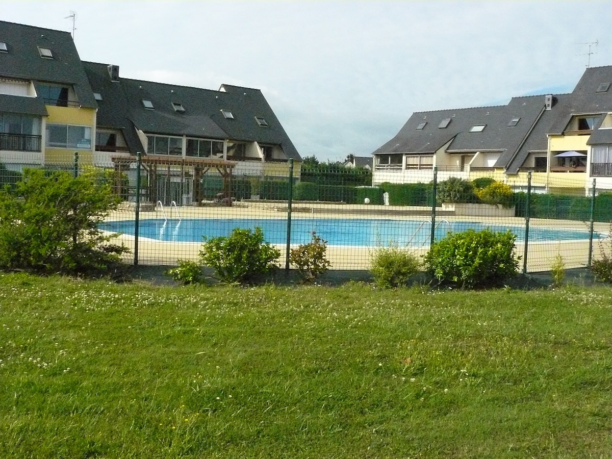 A well-maintained outdoor pool is set within a fenced area, surrounded by green grass and low shrubbery. The structure of nearby residences with sloped roofs and balconies is visible in the background, offering a glimpse of the well-kept environment.