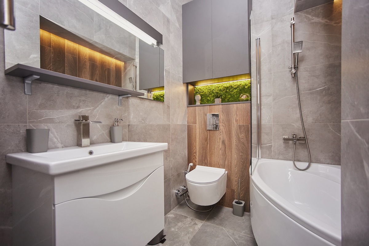 A modern bathroom is displayed, featuring a sleek white vanity with a smooth countertop and a large mirror above. A gray tiled wall contrasts with warm wood accents, and a stylish bathtub is positioned next to a walk-in shower. Subtle lighting enhances the contemporary design.