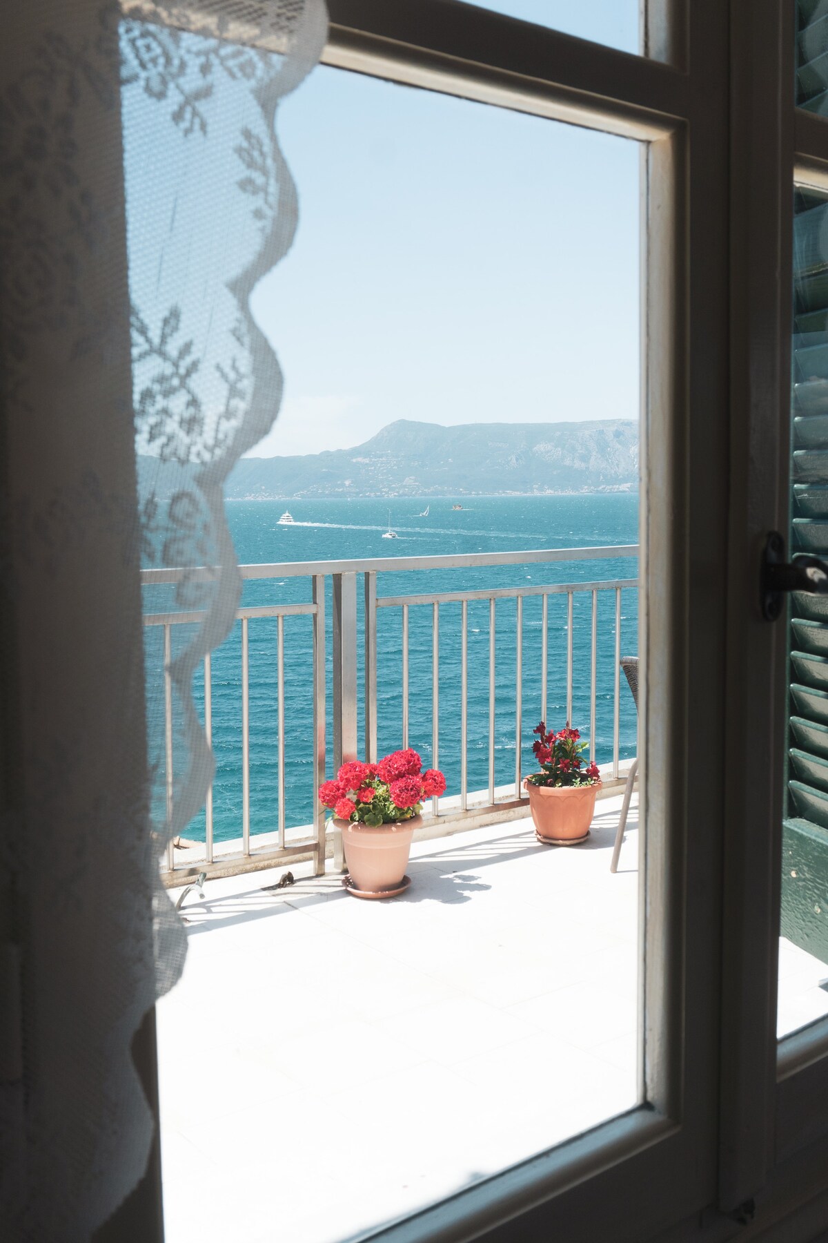 A view is offered from a window, showcasing a balcony with potted red flowers. In the background, the blue sea stretches toward a distant mountainous coastline under a clear sky.