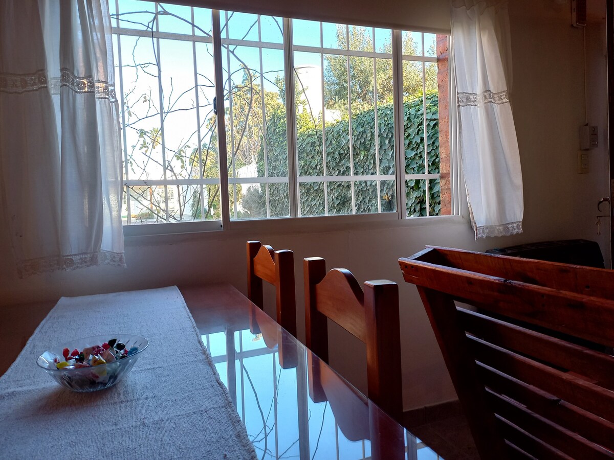 A dining area is visible, featuring a wooden table paired with several matching chairs. Sunlight filters through large windows adorned with sheer white curtains, illuminating the space. Outside, greenery can be glimpsed, creating a connection between indoor and outdoor settings.