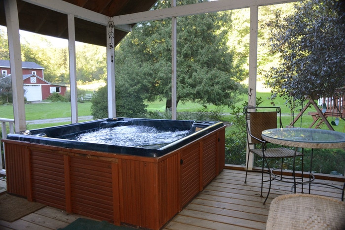 A screened-in porch features a large hot tub, surrounded by wooden decking. A table and chair set is positioned nearby, with lush green trees visible beyond the porch. Natural light filters in, creating a bright and inviting space.