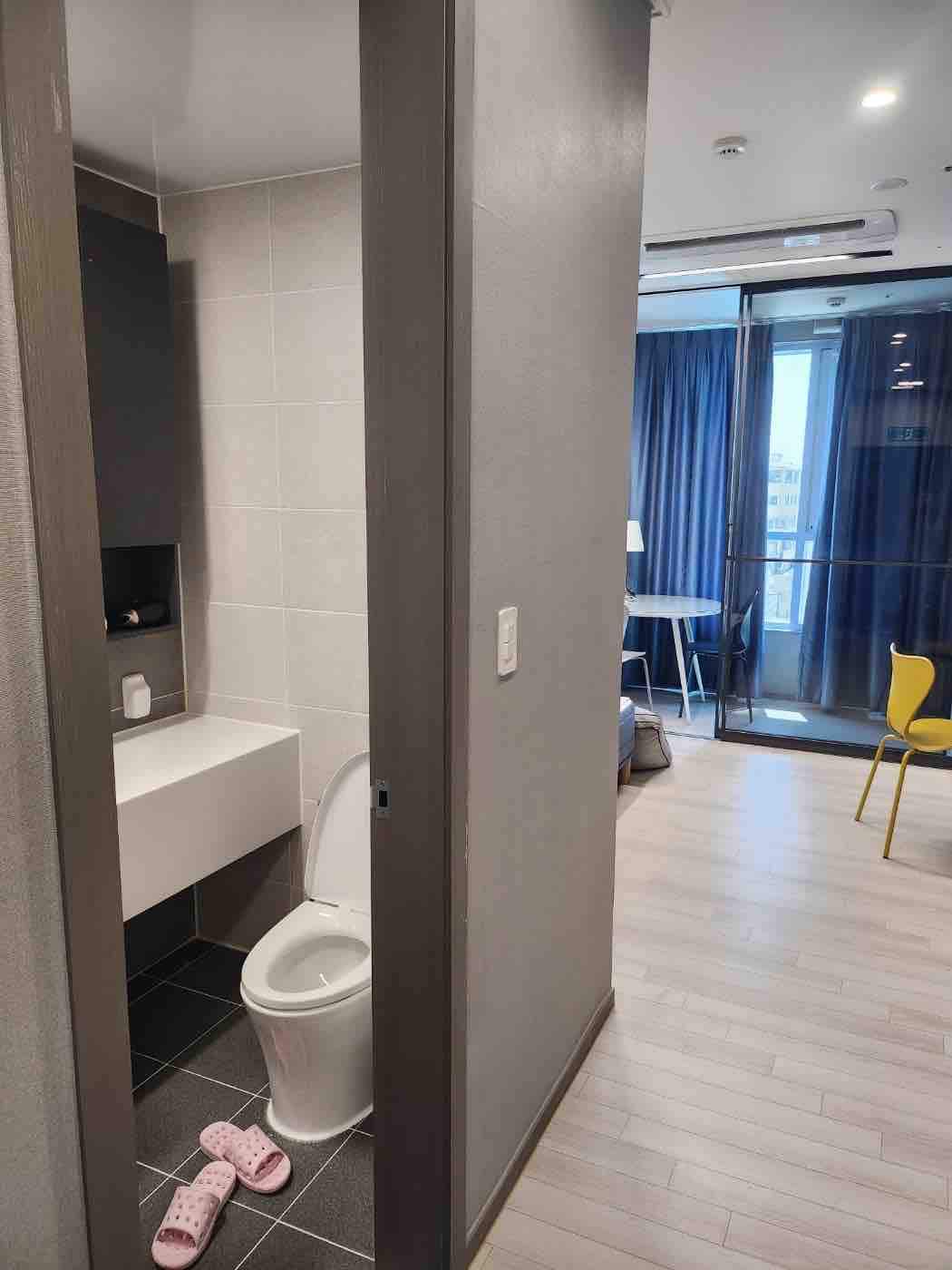 A modern bathroom area is visible, featuring a toilet and a sleek sink. Neutral-tone tiles complement the space. In the background, a well-lit living area can be seen through glass doors, with a dining table and a yellow chair partially visible.
