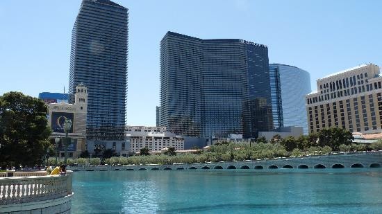 The image captures a view of the Las Vegas Strip, featuring distinctively designed high-rise buildings. The tranquil blue water reflects the structures, while a landscaped area with greenery lies in the foreground. The scene is framed by architectural details and a curved bridge over the water.