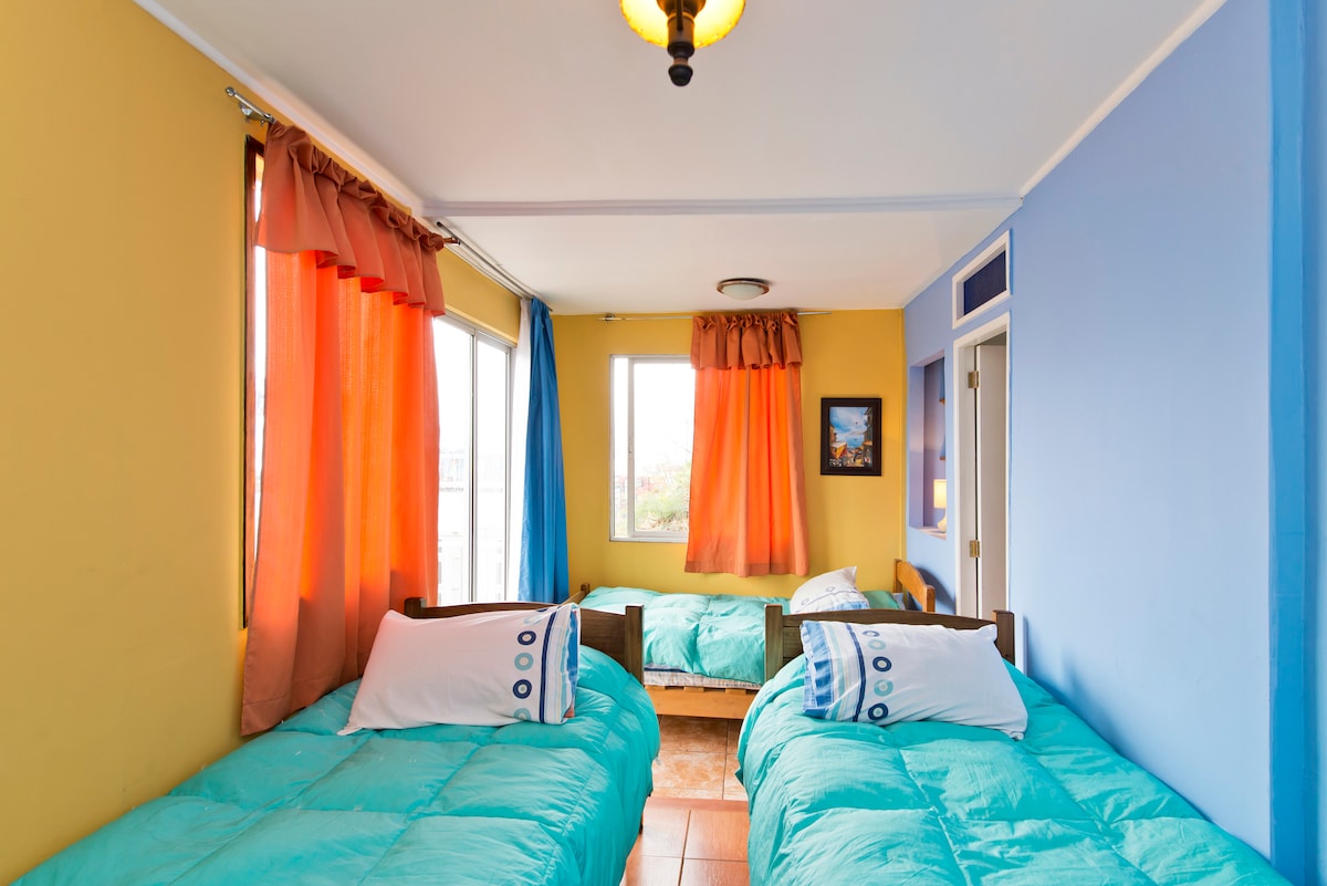 A bright room features three single beds with turquoise bedding. The walls are painted in vibrant yellow and blue, while large windows are dressed with colorful curtains that allow natural light to fill the space.