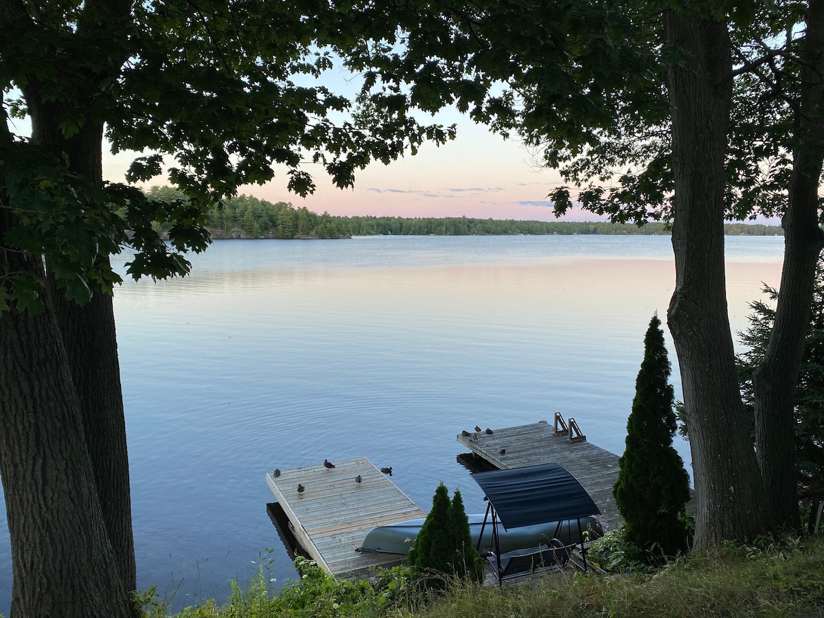 A serene lake view is framed by trees, with calm waters reflecting soft hues of blue and pink. Two private docks are visible along the shoreline, providing direct access to the water. Lush greenery surrounds the area, enhancing the tranquil atmosphere.