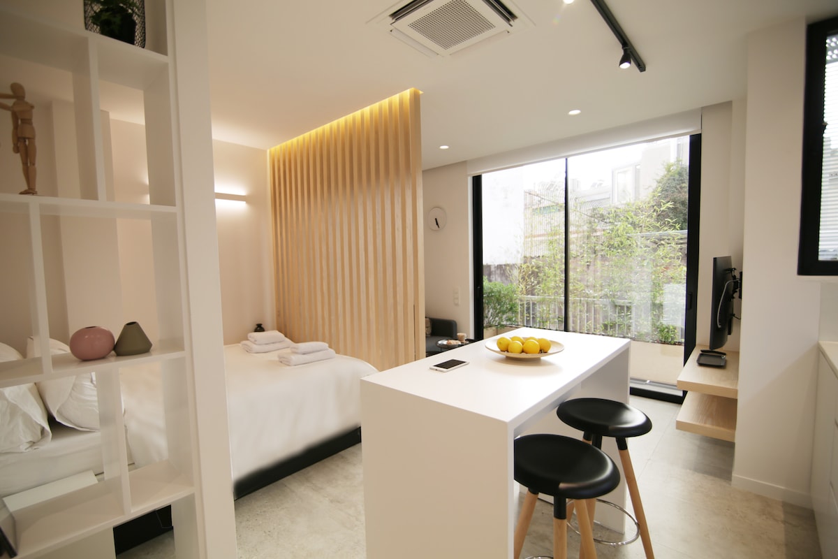 The studio apartment features a minimalistic design with a spacious bed and a small dining area. A large window offers natural light, showcasing greenery outside. The kitchen area includes a bar counter with three stools, complementing the modern aesthetic of the space.