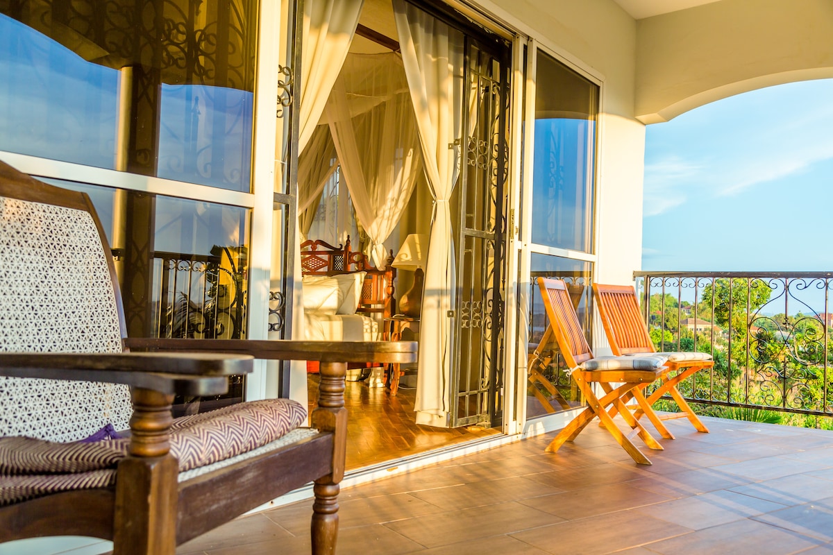 A spacious balcony is furnished with two wooden chairs and offers views of the surrounding landscape. Large glass doors lead into a well-lit interior space adorned with light drapes. The warm wooden flooring complements the inviting atmosphere of the area.