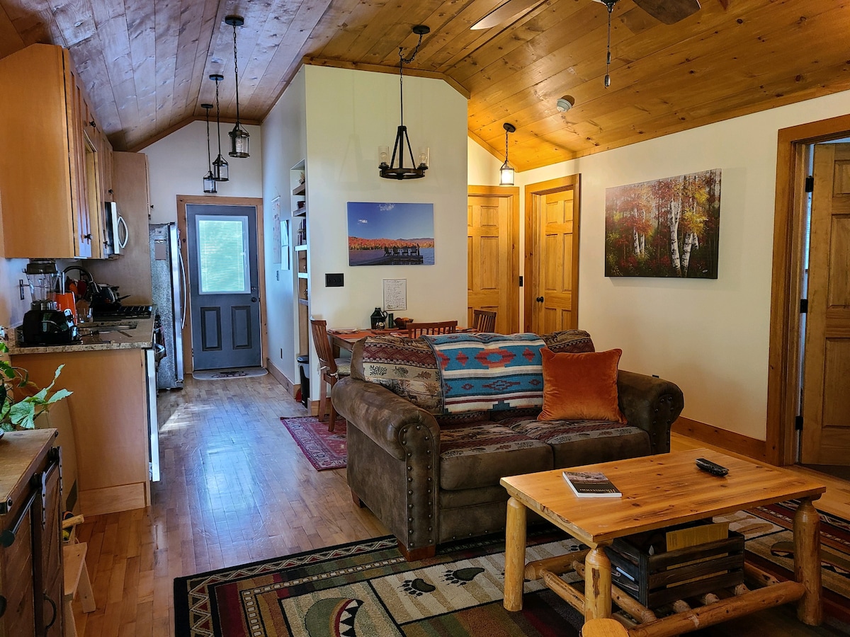 The open living area features a cozy couch adorned with colorful pillows, with a rustic coffee table in front. A wood dining table is visible nearby. Light filters through multiple windows, highlighting wooden ceiling beams and a mix of warm wood and earth-toned furnishings.