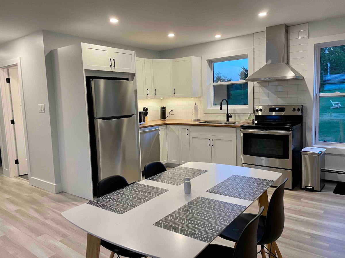 A modern kitchen is featured, equipped with stainless steel appliances, including a refrigerator and a stove. The space includes a sleek dining table surrounded by black chairs, complemented by white cabinetry and light-colored flooring. Natural light is provided through a nearby window.