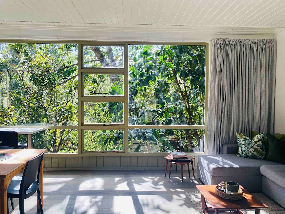 Large windows frame the inviting view of a lush garden filled with greenery. Natural light brightens the lounge area, highlighting the cozy seating arrangement and a small dining table. Soft curtains add a touch of warmth to the serene atmosphere.