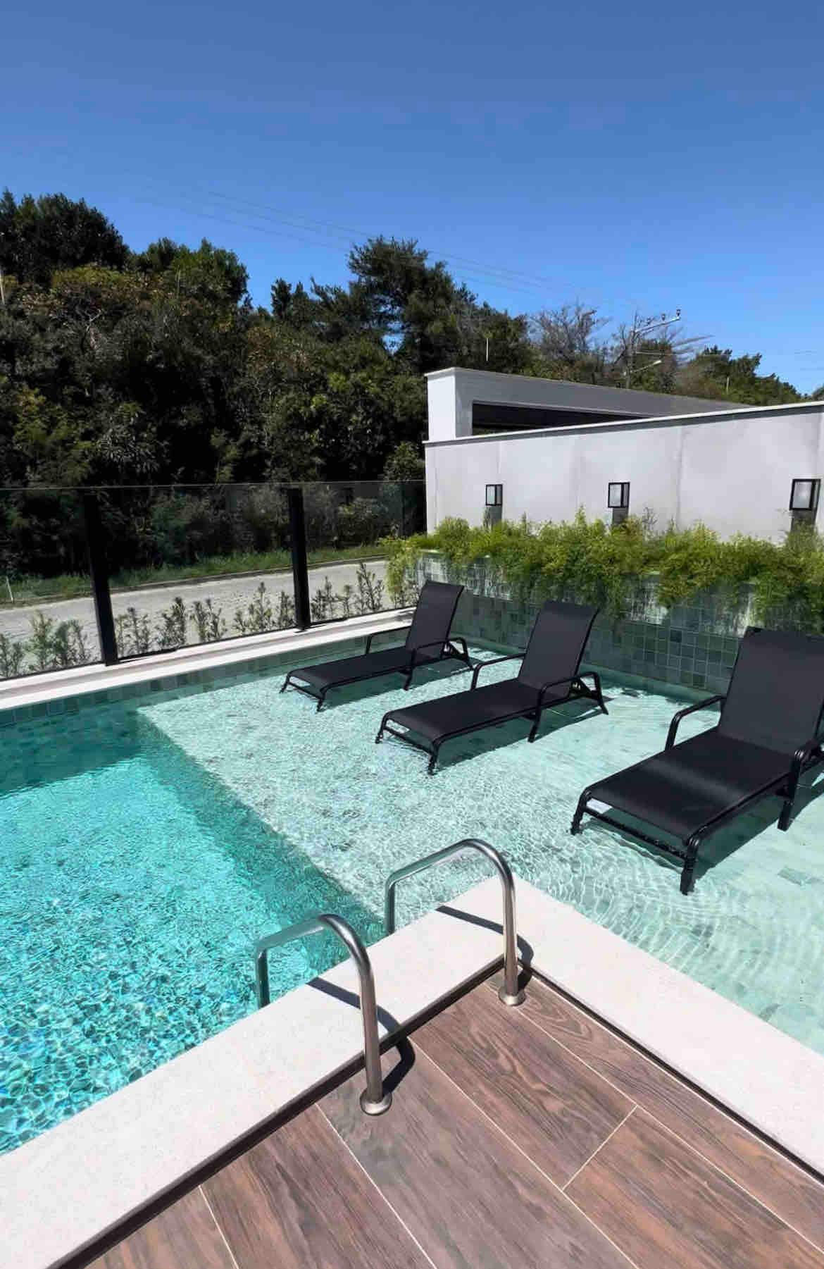 An inviting pool area is visible, featuring crystal-clear water and sleek black lounge chairs positioned around its edge. Lush greenery is observed in the background, while a clear blue sky completes the serene outdoor environment.