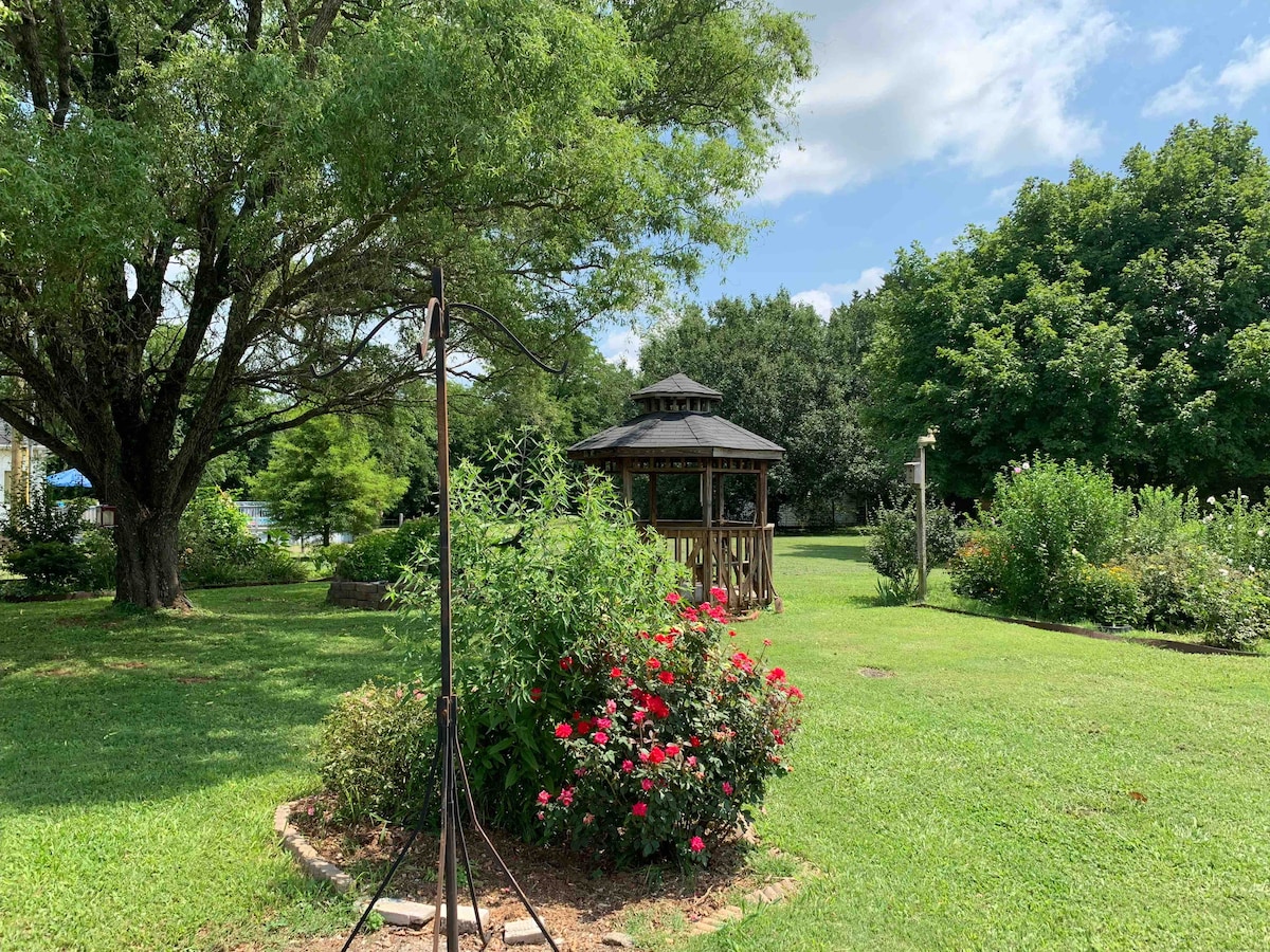 A spacious backyard is adorned with vibrant flower beds, including blooming red roses. A gazebo is positioned centrally, surrounded by lush greenery and large trees, providing shade. The open lawn invites relaxation and leisure in a serene outdoor environment.