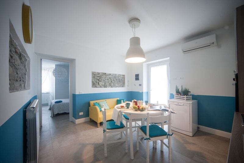A bright dining area features a white table set with colorful dishware and fresh fruit. Blue and white walls create a modern ambiance, complemented by a comfortable yellow sofa. An air conditioning unit is mounted above, and an open doorway provides a glimpse into a nearby bedroom.