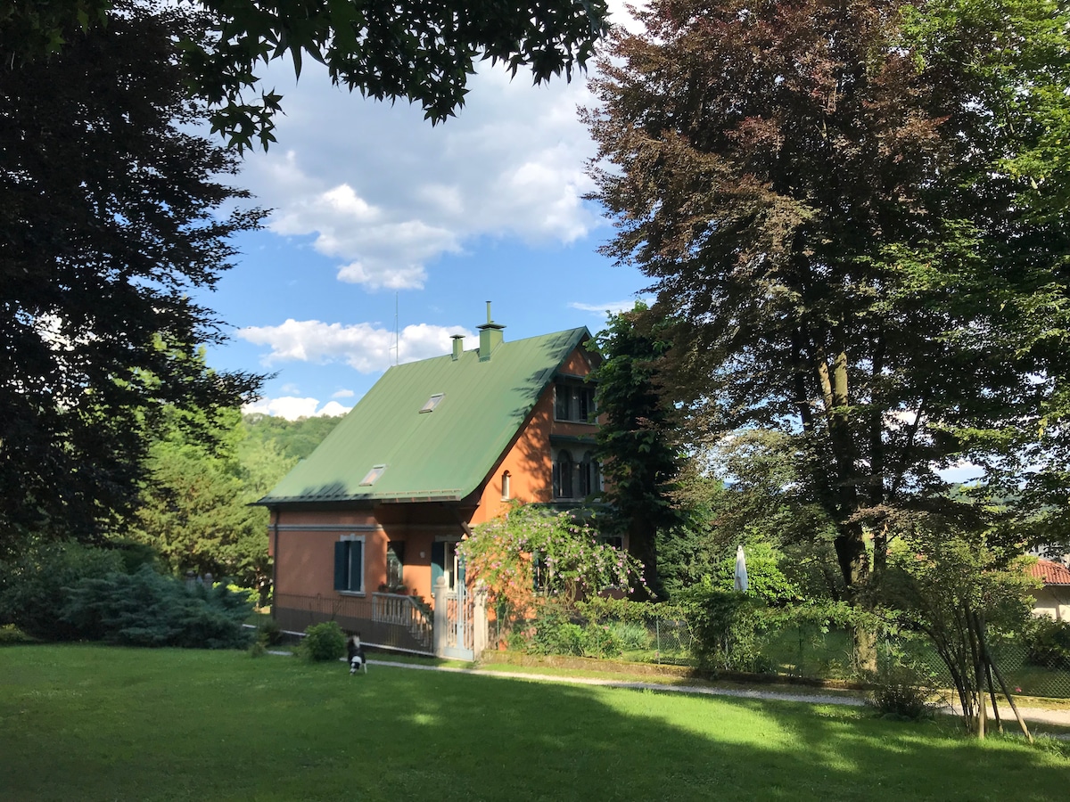 A three-story villa is set amidst lush greenery, featuring a green roof and earthy-toned exterior. The surrounding garden includes tall trees and well-maintained grass, giving a serene and private atmosphere. A small balcony can be seen on the front of the house.