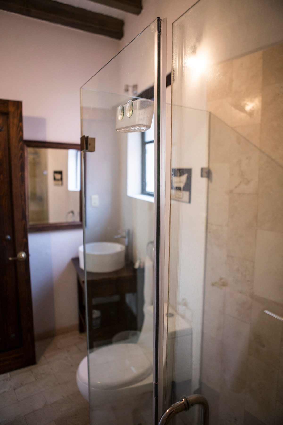 A contemporary bathroom features a glass shower enclosure, allowing natural light to fill the space. The room includes a modern toilet and a circular sink atop a wooden vanity. A mirror is positioned above the sink, complementing the neutral tiled walls.