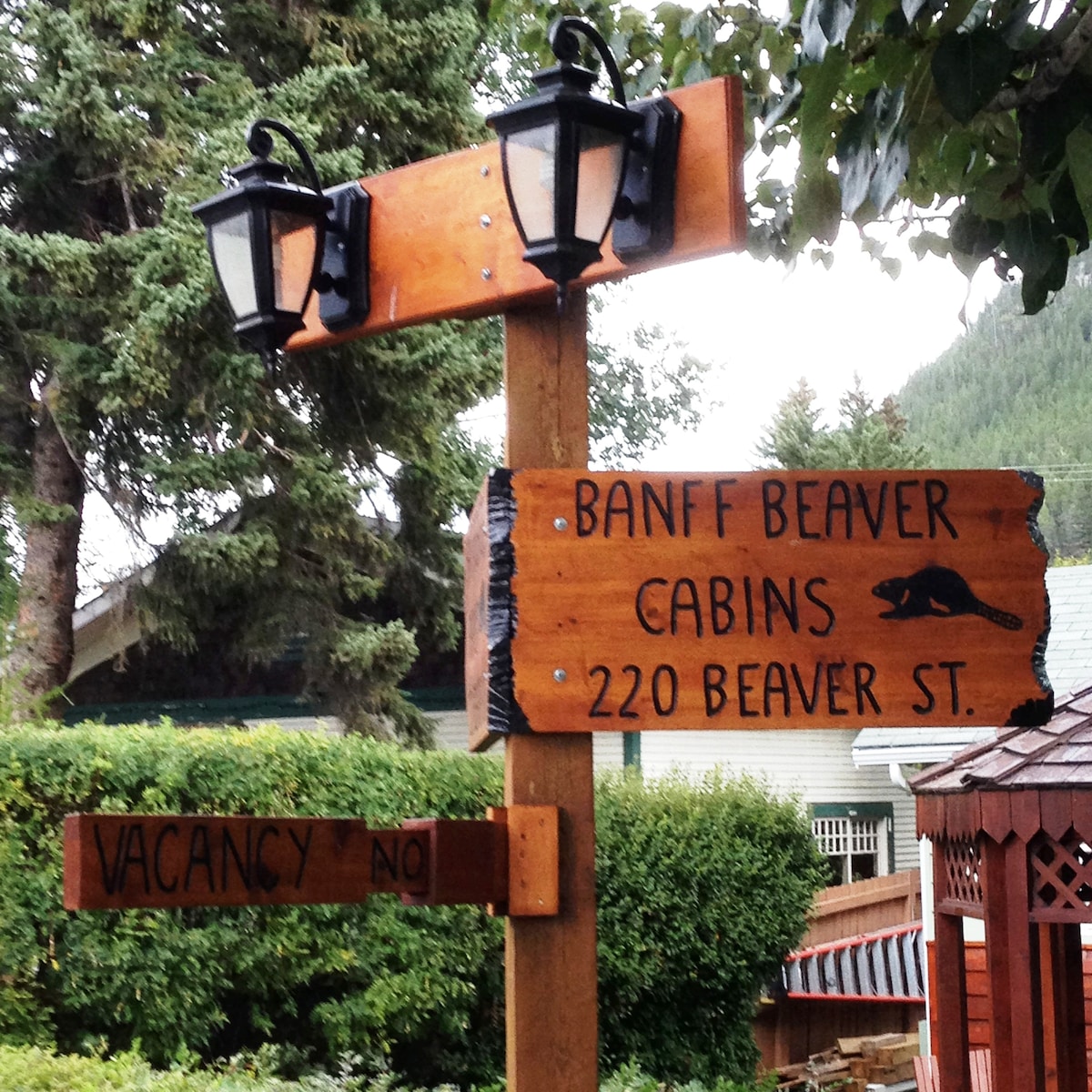 A wooden signpost displays the name 'Banff Beaver Cabins' and the address '220 Beaver St.' Two lantern-style lights are mounted above the sign, providing illumination. A second sign below indicates 'VACANCY NO.' Surrounding greenery and a gazebo contribute to a natural, inviting setting.