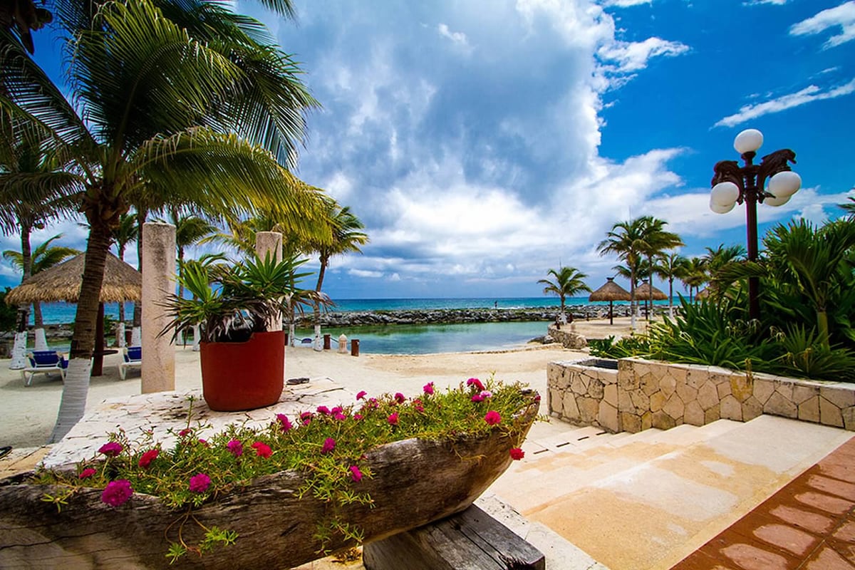 A serene beachfront area is framed by palm trees and vibrant flowers. The sandy shore leads to clear waters, while thatched umbrellas and lounge chairs are arranged nearby. A rocky jetty and distant horizon under a cloudy sky are part of the scenic view.