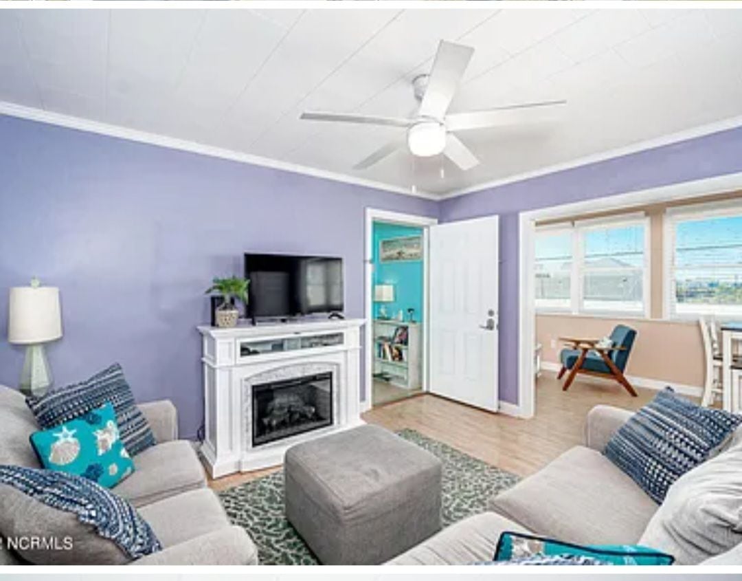 A light-filled living space features a lavender accent wall, a ceiling fan, and a cozy seating arrangement. A television is mounted above a decorative fireplace. A sunroom is visible through an open door, showcasing additional seating and natural light.