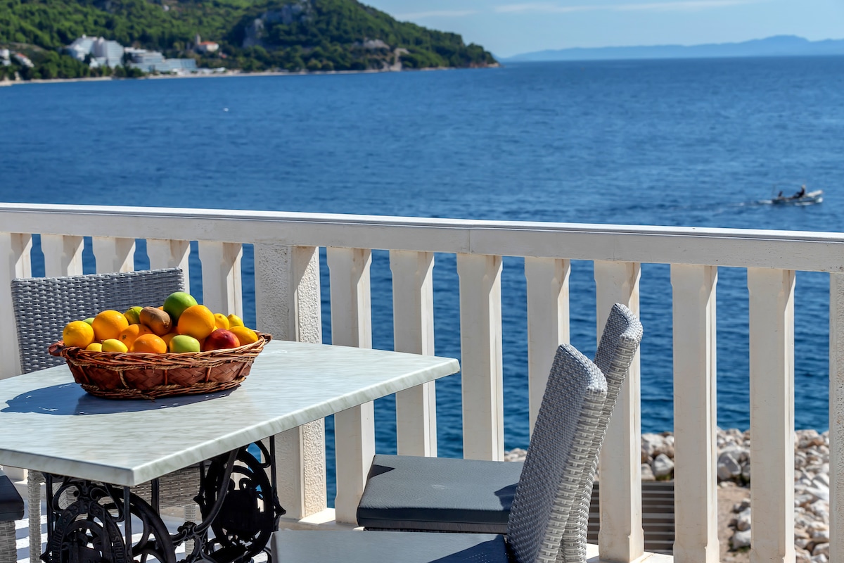 A dining table with a marble top is set on a sunny terrace, featuring a wicker basket filled with colorful fruits. The view includes clear blue waters and distant hills, emphasizing a serene coastal setting.