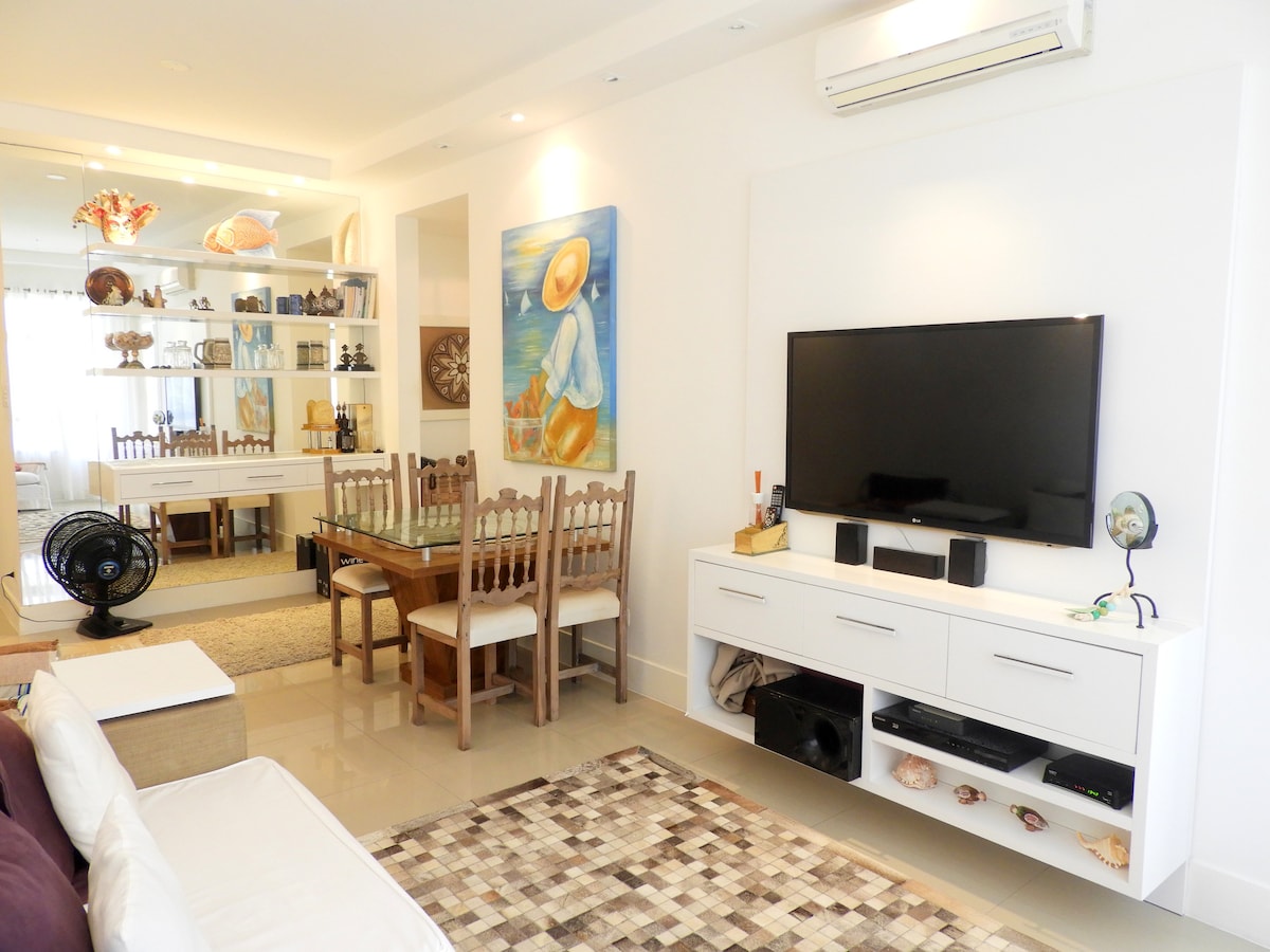 The living space features a mounted television flanked by a modern entertainment unit. A dining table with four chairs is positioned nearby. An air conditioning unit is installed overhead, and decorative elements, including artwork and shelves displaying various items, contribute to the room's ambiance.