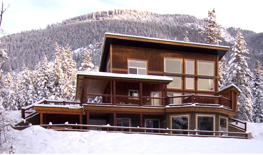 A spacious chalet is nestled in a snowy landscape, featuring large windows that provide views of the surrounding mountains. The structure showcases multiple levels and an expansive deck, ideal for enjoying the outdoor scenery. The exterior is adorned with natural wooden elements.