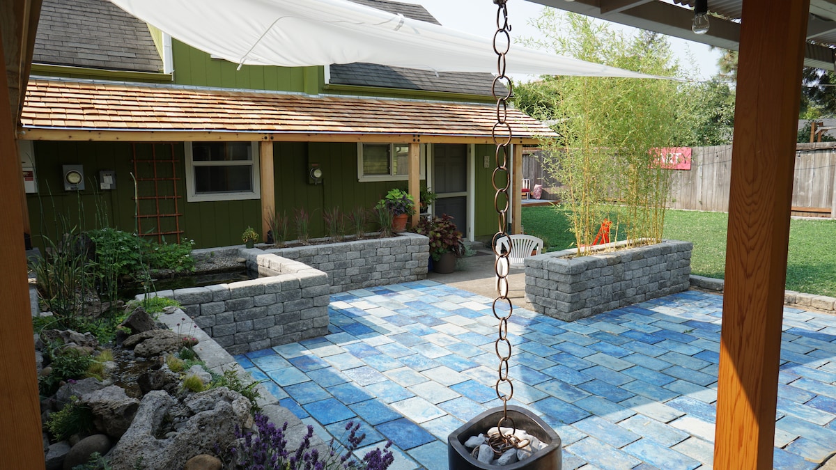 An outdoor garden area features a stone patio laid with blue tiles, bordered by low stone planters filled with plants. A relaxing space is complemented by a hanging chain connected to a small fire pit, with greenery and the structure in the background.