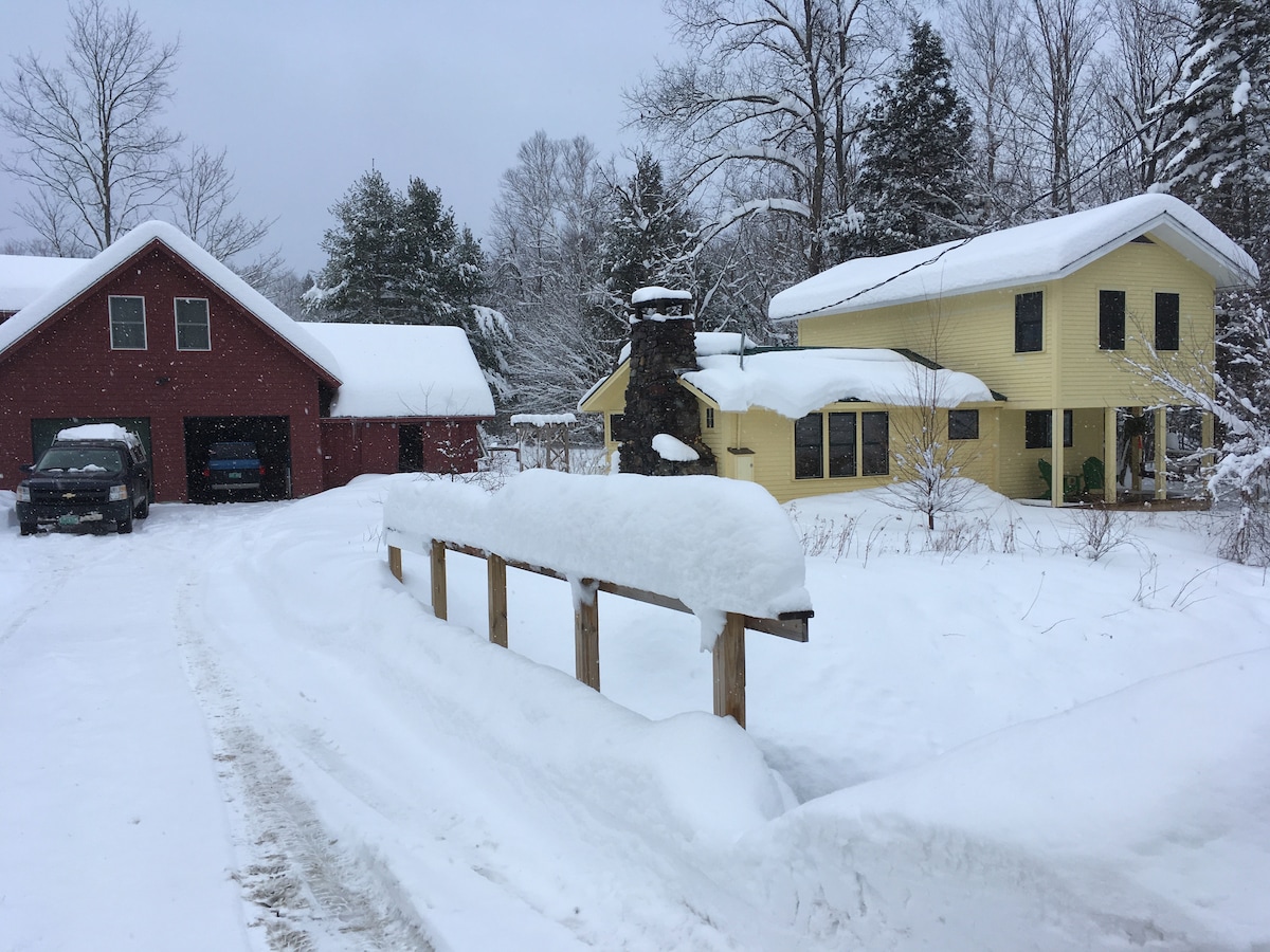 A snow-covered landscape features a yellow house and a red garage. The driveway is lined with snow, creating a serene winter scene. Leafless trees are visible in the background, and snow is accumulating on the roofs, enhancing the tranquil atmosphere of the rural setting.