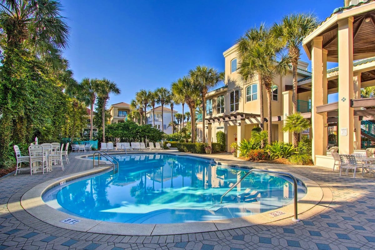 A resort-style pool is set against a backdrop of palm trees and landscaped greenery. Lounge chairs surround the pool area, and a shaded pavilion is visible, enhancing the space for relaxation. The clear water reflects the blue sky, inviting a refreshing dip.
