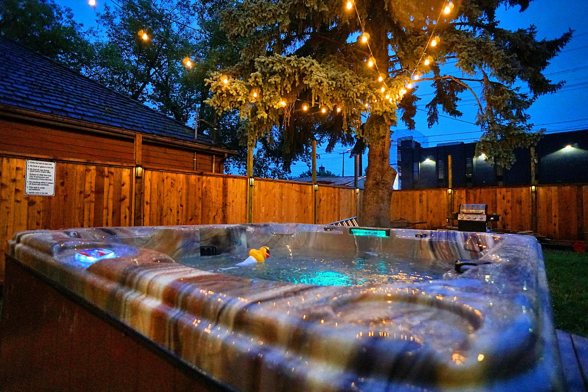 A hot tub sits in a well-lit backyard, surrounded by wooden fencing. Soft glow from string lights above adds to the ambiance. A tree stands nearby, with a rubber duck floating in the water, inviting relaxation under the evening sky.