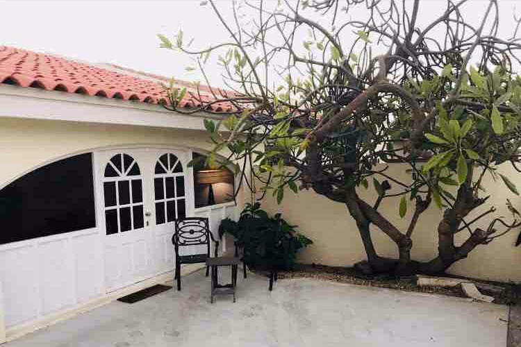 An outdoor patio area features a small table with two chairs, shaded by a large tree. Nearby, a white door with multiple arches leads into the interior space, creating a welcoming entryway.