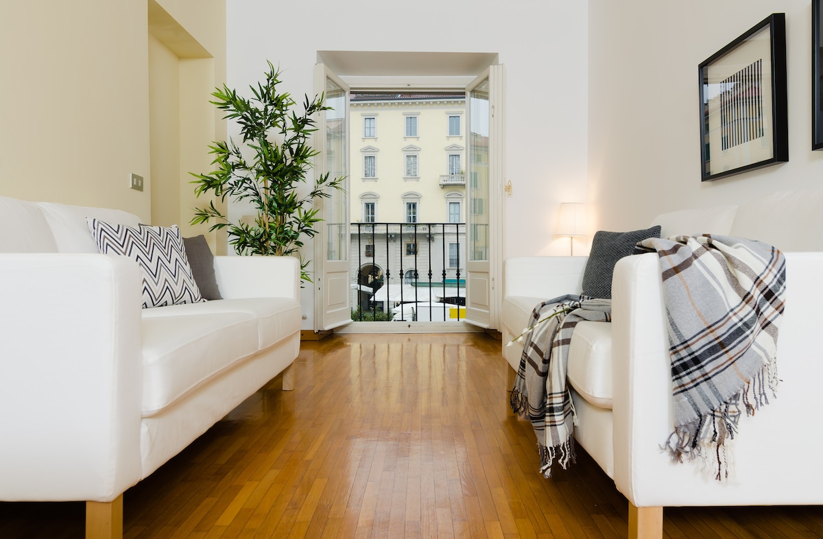A bright living area features two white sofas arranged for relaxation. A large window opens to the street, allowing natural light to fill the space. A potted plant adds a touch of greenery, while a throw blanket is draped over one sofa.