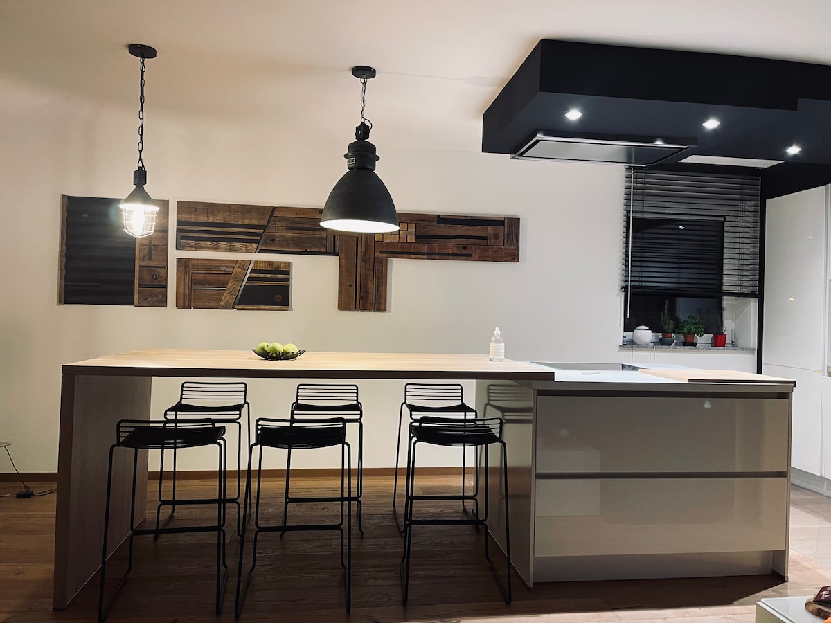 A modern kitchen features a central island with four sleek bar stools arranged around it. Ambient lighting is provided by two pendant lamps above the island, while wooden wall art adds a touch of texture to the bright space.