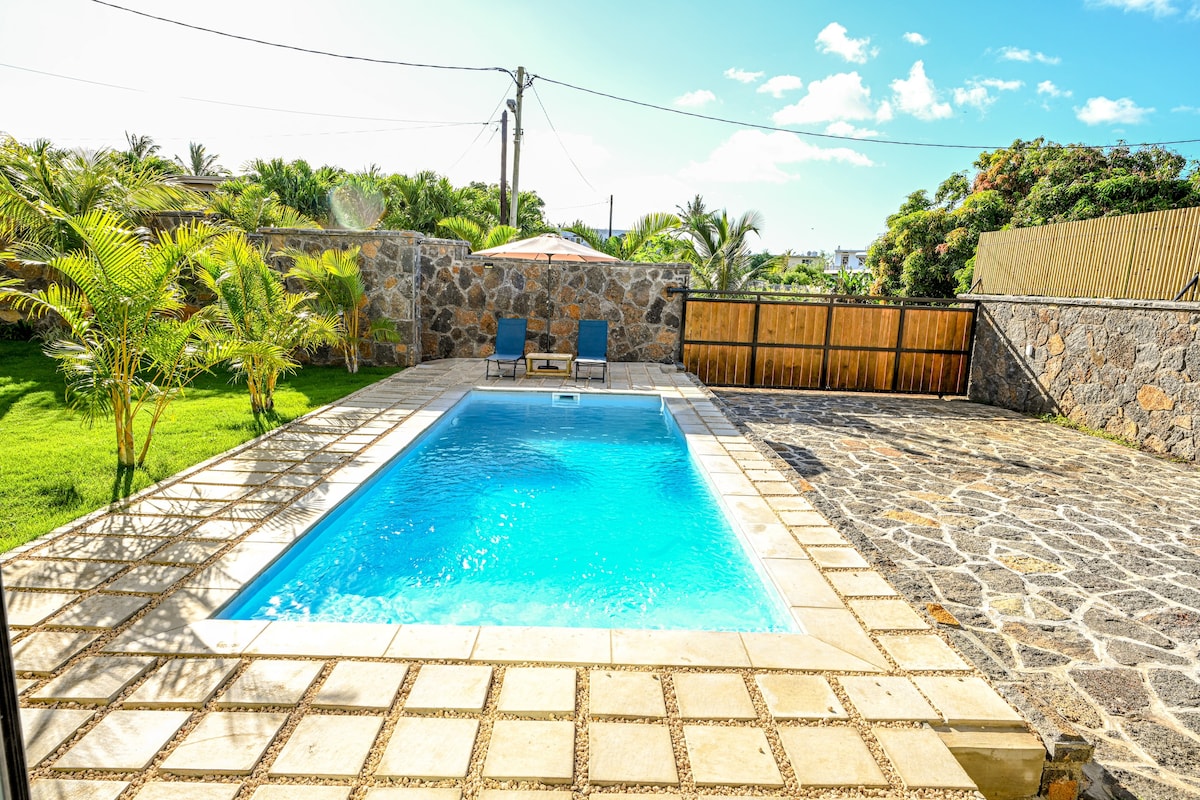 A private swimming pool is framed by a spacious stone patio and lush greenery, offering a serene outdoor setting. Two lounge chairs are positioned by the pool, inviting relaxation under the clear blue sky. The fenced area ensures a sense of privacy and security.