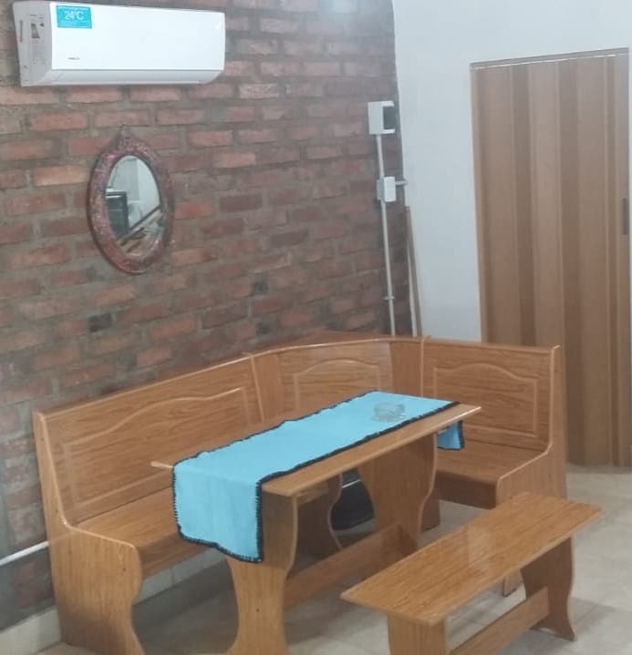 A wooden dining set is positioned against a textured brick wall. The set includes a table with a light blue runner and a corner bench. A mirror hangs above the table, while an air conditioning unit is mounted on the wall.