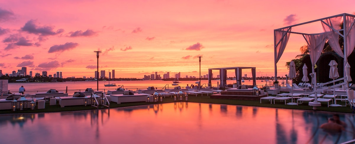 A serene view of a swimming pool reflects a vibrant sunset sky, with hues of pink and orange illuminating the horizon. Lounge chairs are arranged around the pool area, while elegant cabanas stand in the background, offering a backdrop of downtown Miami's skyline.