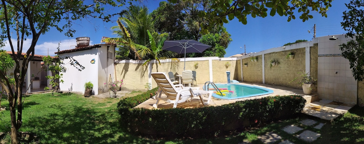 A well-maintained outdoor area features a swimming pool surrounded by lush greenery and a sunbathing space with lounge chairs. An umbrella provides shade, while a small structure is visible in the background. The tranquil environment is enhanced by soft landscaping elements.