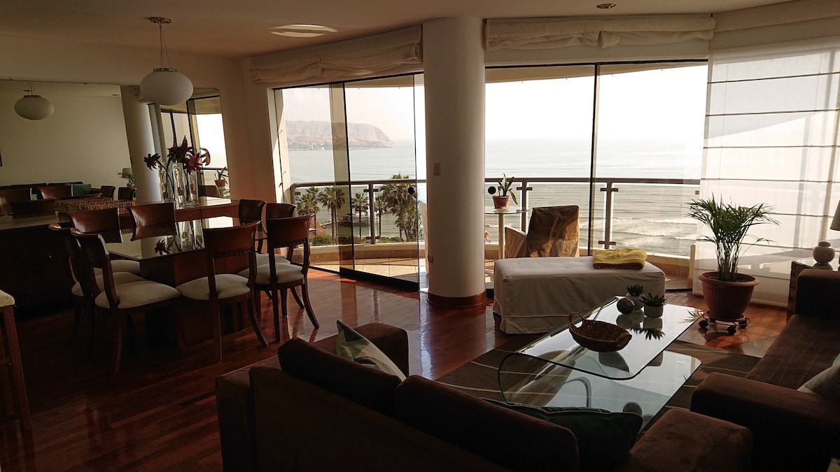 A spacious living area is filled with natural light, featuring large windows with views of the sea. A comfortable sofa is complemented by a glass coffee table, while a dining table is set for six. Potted plants add a touch of greenery to the room.