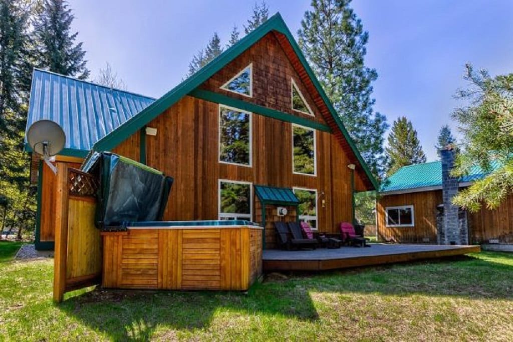 A spacious cabin features a striking A-frame design with a textured wooden exterior and a vibrant teal roof. A private hot tub is situated on a wooden deck within a grassy area, surrounded by lush trees, enhancing the tranquil outdoor setting.