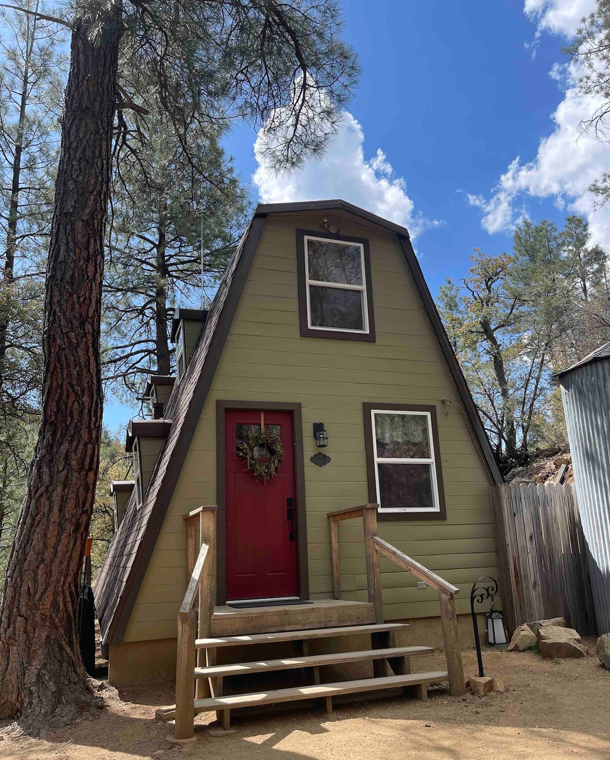 A triangular-shaped cabin is shown, featuring green siding and a red door. Steps lead up to the entrance, framed by a wooden railing. Tall ponderosa pines are visible nearby, with a blue sky and white clouds above, creating a serene outdoor setting.