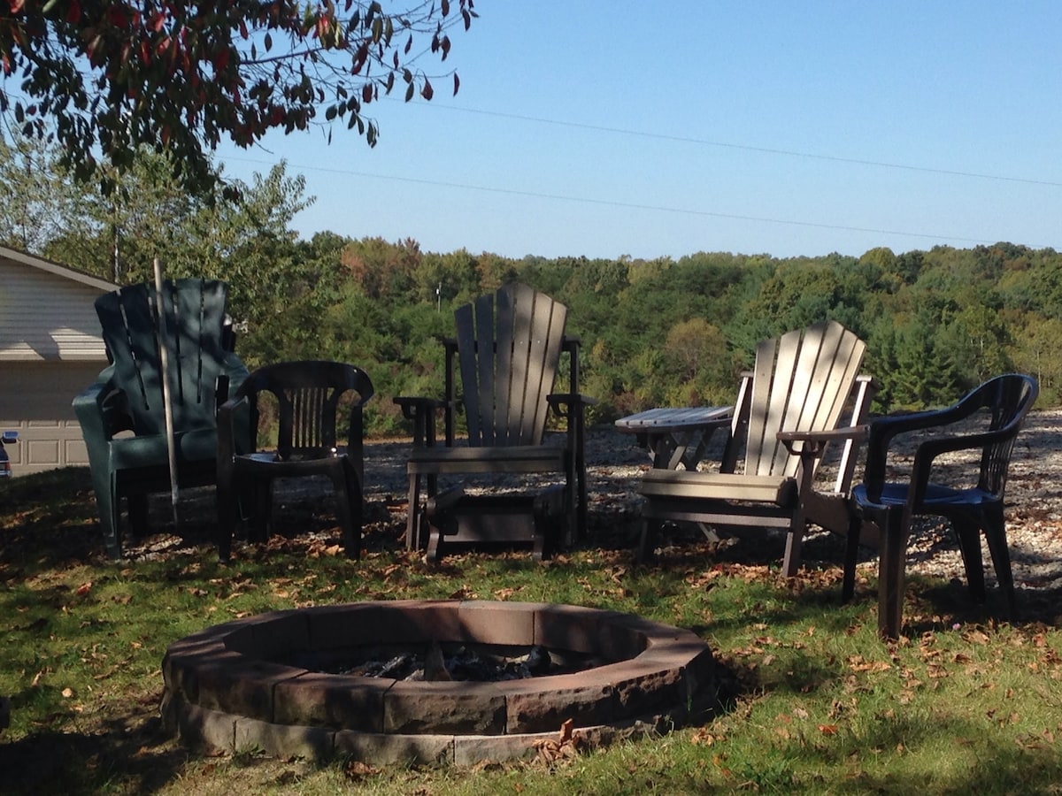 A fire pit surrounded by a variety of seating options, including wooden Adirondack chairs and plastic lawn chairs, is situated on a grassy area. The scenic backdrop features rolling hills with trees that exhibit fall colors, under a clear blue sky.