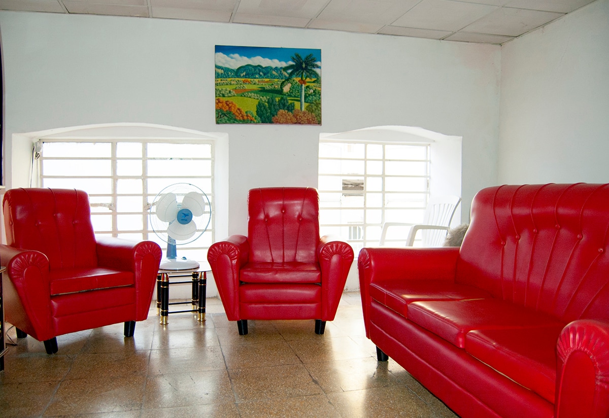 A bright living area is furnished with red leather sofas, creating a warm seating arrangement. A coffee table sits between the sofas. Light streams in through two large windows, complemented by a blue fan for ventilation and a landscape painting on the wall.