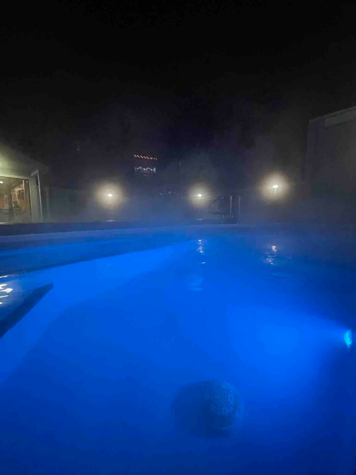 A heated pool is illuminated in soft blue light, creating a serene atmosphere. Mist rises softly from the surface, while dim lights surrounding the area enhance the tranquil setting. The image suggests a peaceful retreat for evening relaxation.