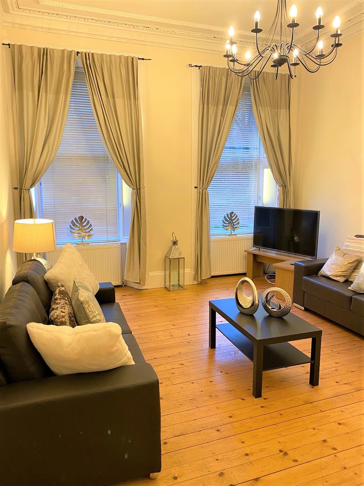 A living area is presented with two sofas and a coffee table situated on warm wooden flooring. Tall windows draped with light curtains allow natural light to enter. A stylish chandelier hangs from the ceiling, and decorative elements are thoughtfully placed throughout the space.