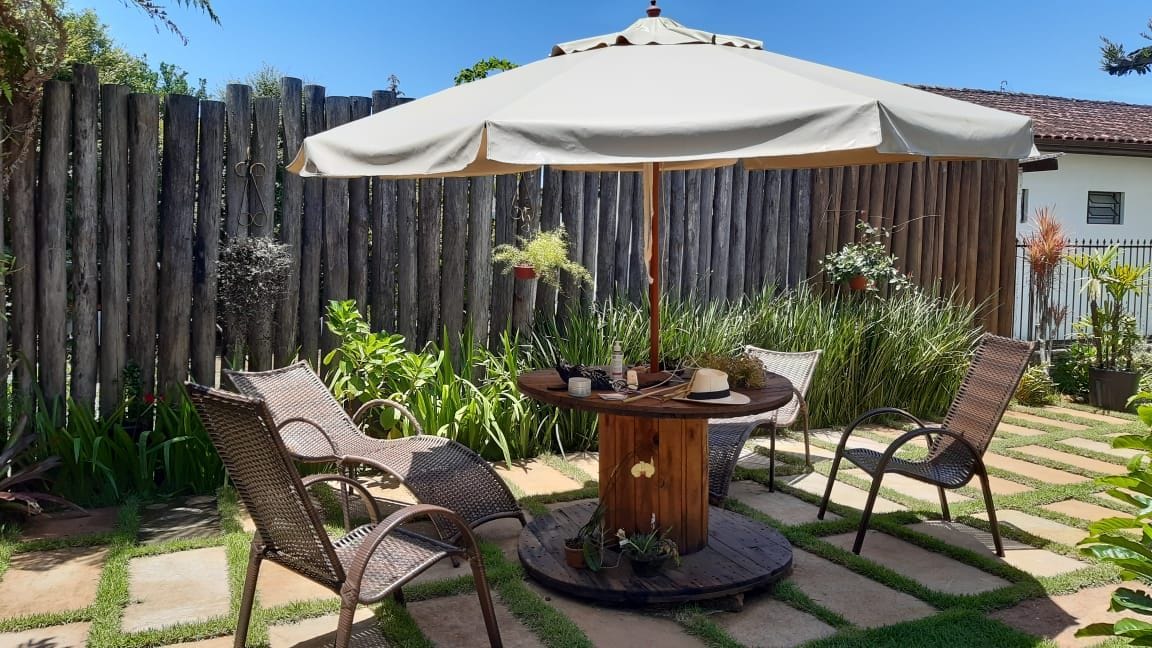 An outdoor seating area is arranged beneath a large umbrella, featuring a round wooden table at its center. Four wicker chairs are positioned around the table, surrounded by lush greenery and a wooden fence in the background.