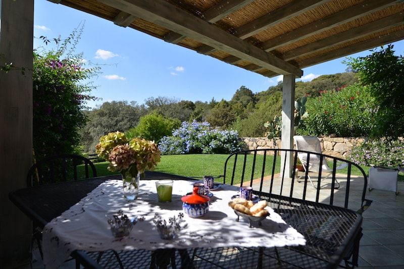A shaded outdoor dining area features a table with black metal chairs. A light tablecloth is draped over the table, which is set with snacks. Lush greenery and flowering plants create a scenic backdrop, complemented by blue skies and gentle sunlight.