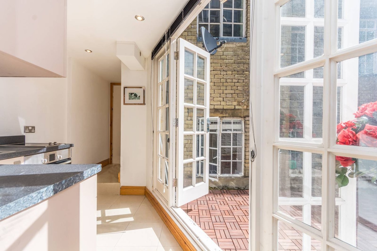 A bright kitchen area features large glass doors that open to a private terrace. A sleek countertop and stove are visible, combining functionality with natural light. The terrace outside showcases a brick pattern and floral arrangements provide a touch of color, enhancing the outdoor view.