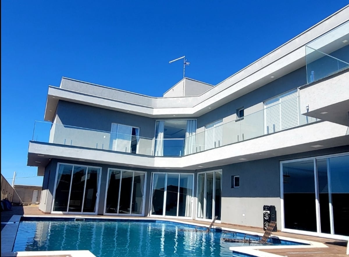 A modern building with multiple levels showcases expansive glass windows and sliding doors, allowing for natural light and views of the outdoor area. The swimming pool reflects the clear blue sky, and a wooden deck surrounds the pool area, providing space for relaxation.