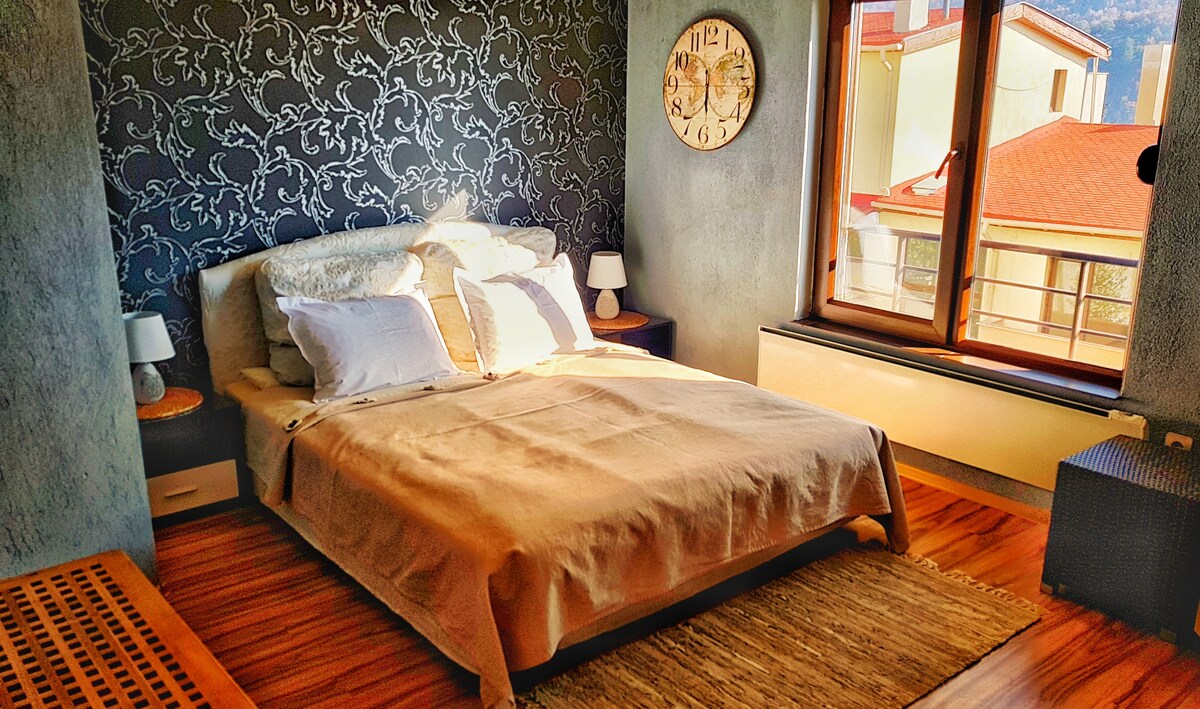 A well-appointed bedroom features a spacious bed with neutral linens, accented by white pillows. Nightstands with lamps are positioned on either side. A large window offers natural light and a view of the outside, framed by decorative wallpaper and wooden flooring.