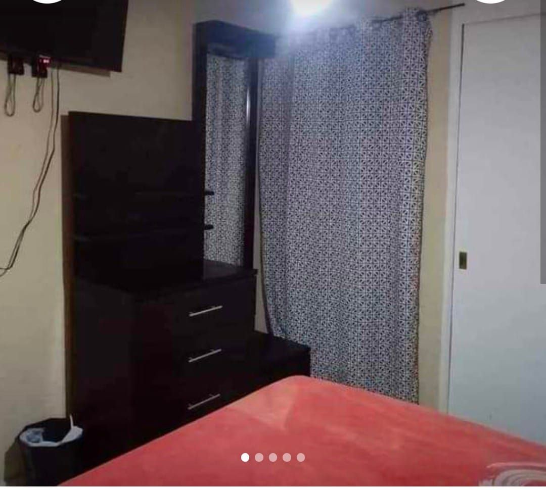 A neatly arranged corner of the room features a dresser with three drawers beside a full-length mirror. Light fabric curtains are mounted on a rod, offering subtle privacy. A wall-mounted television is positioned for viewing convenience, and a wastebasket is present on the floor.